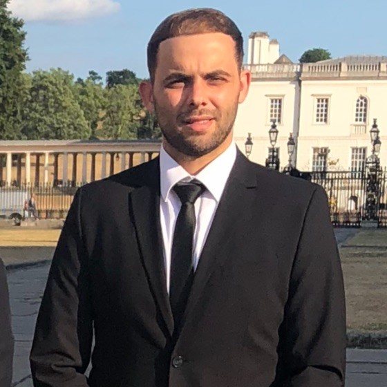 A man in a black suit and tie stands outdoors in front of a white historic building on a sunny day, attending the Memcyco at 2nd Edition Fraud & Scams Leaders Summit.
